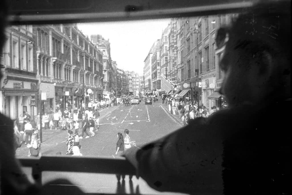 Plongée sur une rue de Londres vue à travers la vitre frontale d'un bus à impériale, encadrée par la silhouette d'un homme de profil.